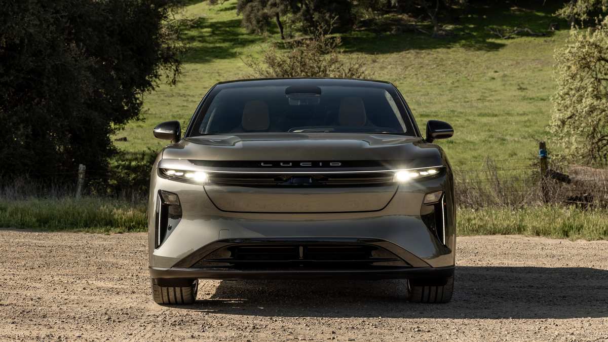 A beige 2025 Lucid Gravity with bold headlights sits on a dirt road against a lush green hillside background, conveying power and modern elegance.