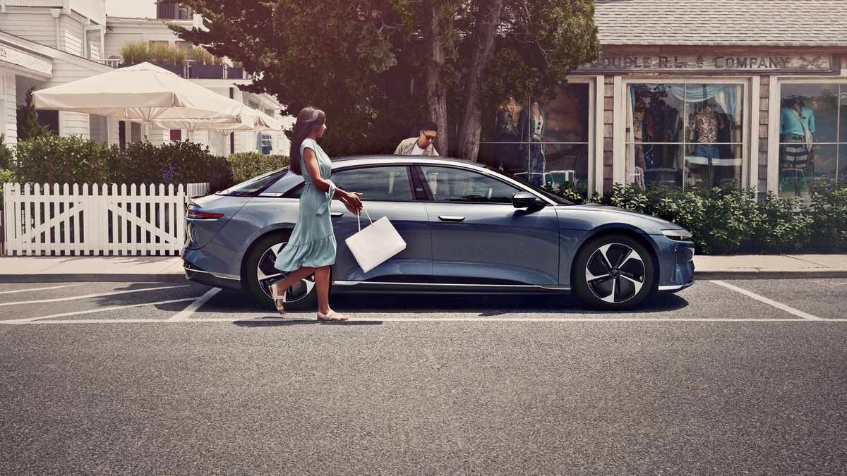 A woman in a light blue dress walks past a sleek, modern car parked on a street lined with greenery and shops in the background.