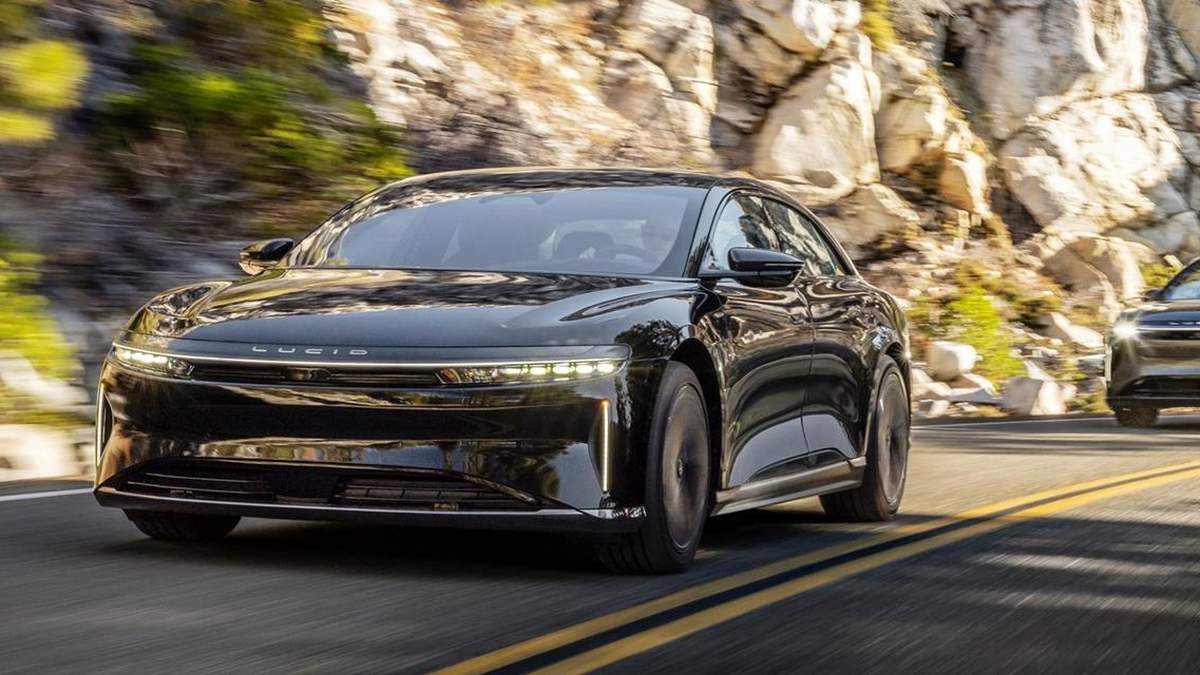 A sleek black Lucid electric car accelerates along a winding mountain road, surrounded by rocky cliffs and greenery.