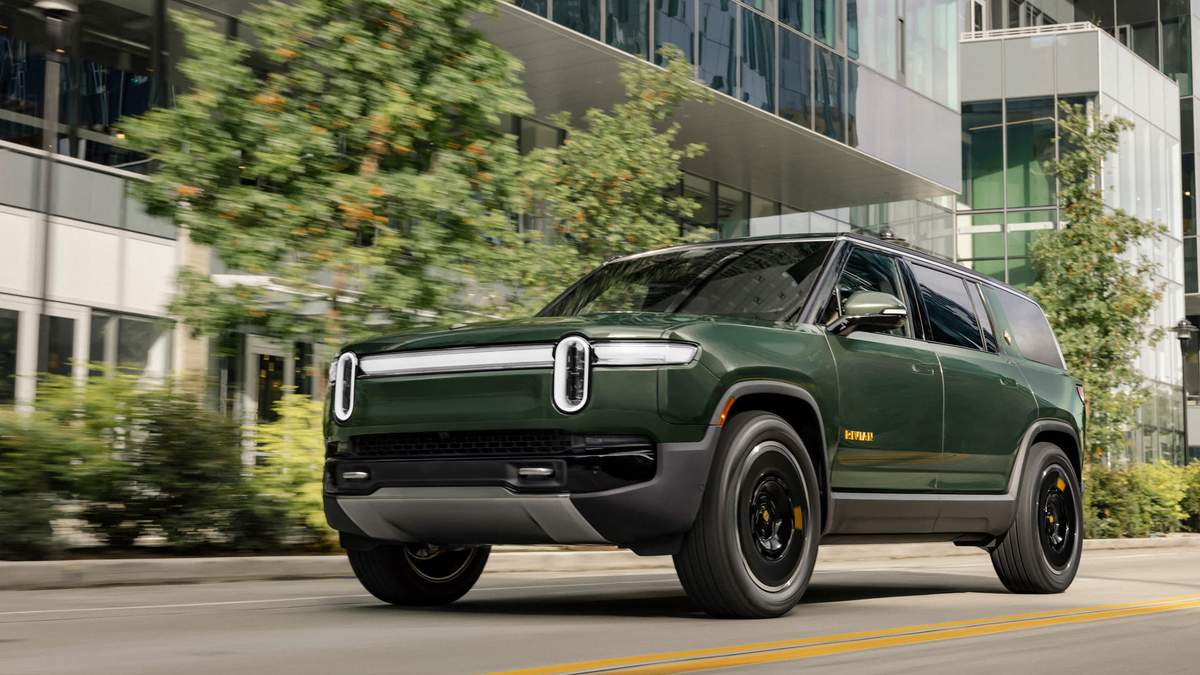 A green 2025 Rivian R1S, featuring modern design and striking headlights, drives through an urban landscape with lush trees.