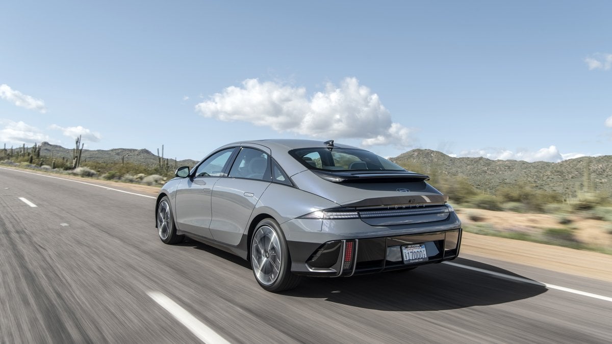 A Silver Hyundai Ioniq 6 accelerates on a desert highway, surrounded by mountains and cacti under a blue sky with fluffy clouds.