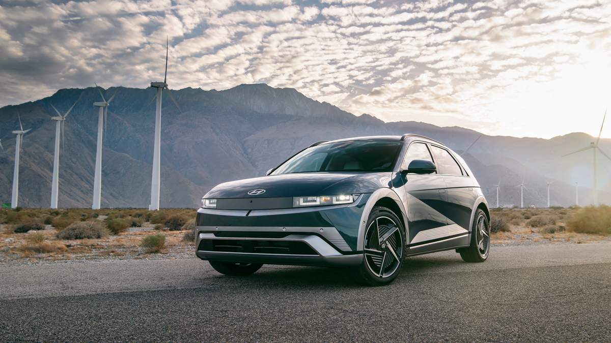 A Hyundai Ioniq 5 parked on a road, with wind turbines and mountains in the background under a cloudy sky.