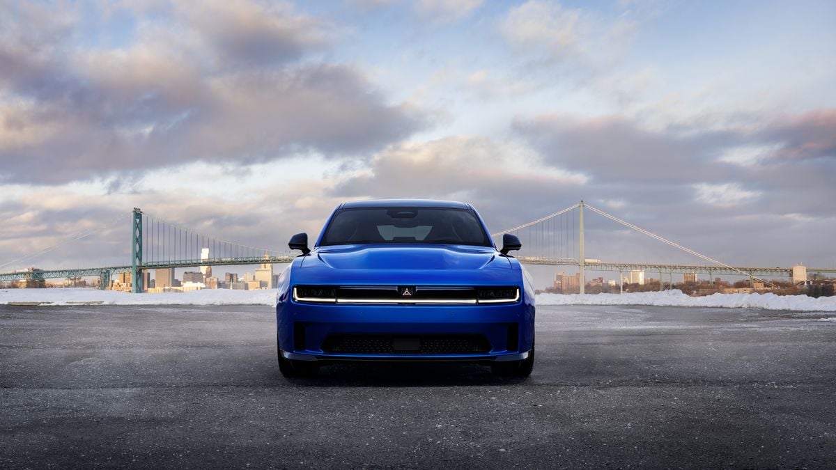 A 2025 Dodge Charger EV is parked in front of a bridge and city skyline under a cloudy sky, showcasing a modern design and urban backdrop.