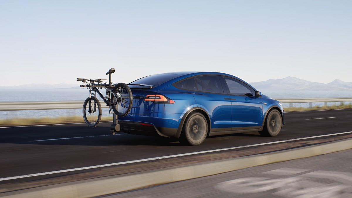 A blue Tesla Model X drives on a road, with a bicycle mounted on a rear rack, against a scenic background of mountains and water.