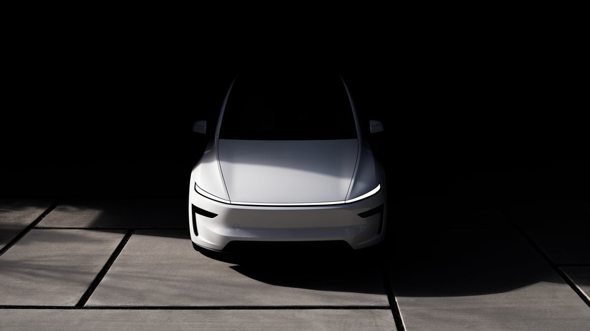 A white 2026 Tesla Model Y Juniper, front view emerging from darkness, illuminated by soft light on a textured concrete surface.