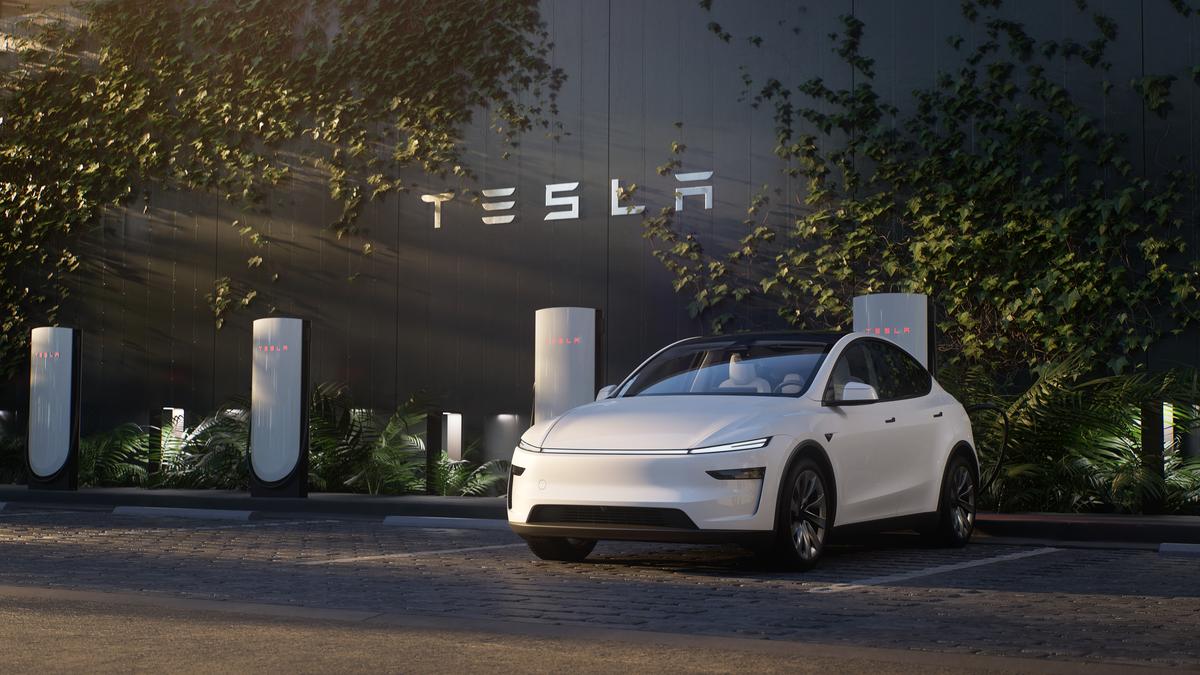 A white 2026 Tesla Model Y Juniper parked in front of a Tesla charging station surrounded by greenery and a black wall with the Tesla logo.
