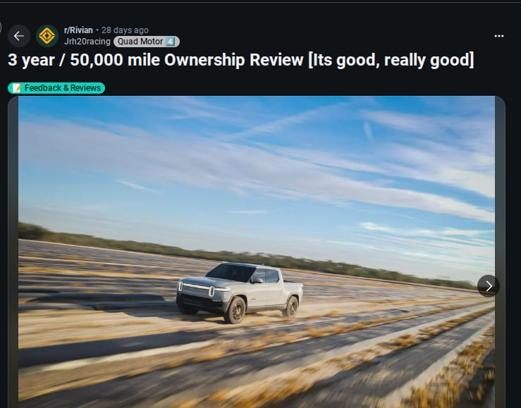 A Rivian electric truck drives swiftly across a rural landscape, showcasing its sleek design against a blue sky.