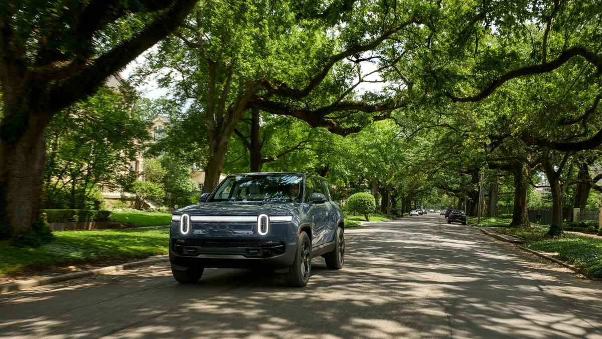 A blue electric SUV drives down a tree-lined street, surrounded by lush greenery on a sunny day.