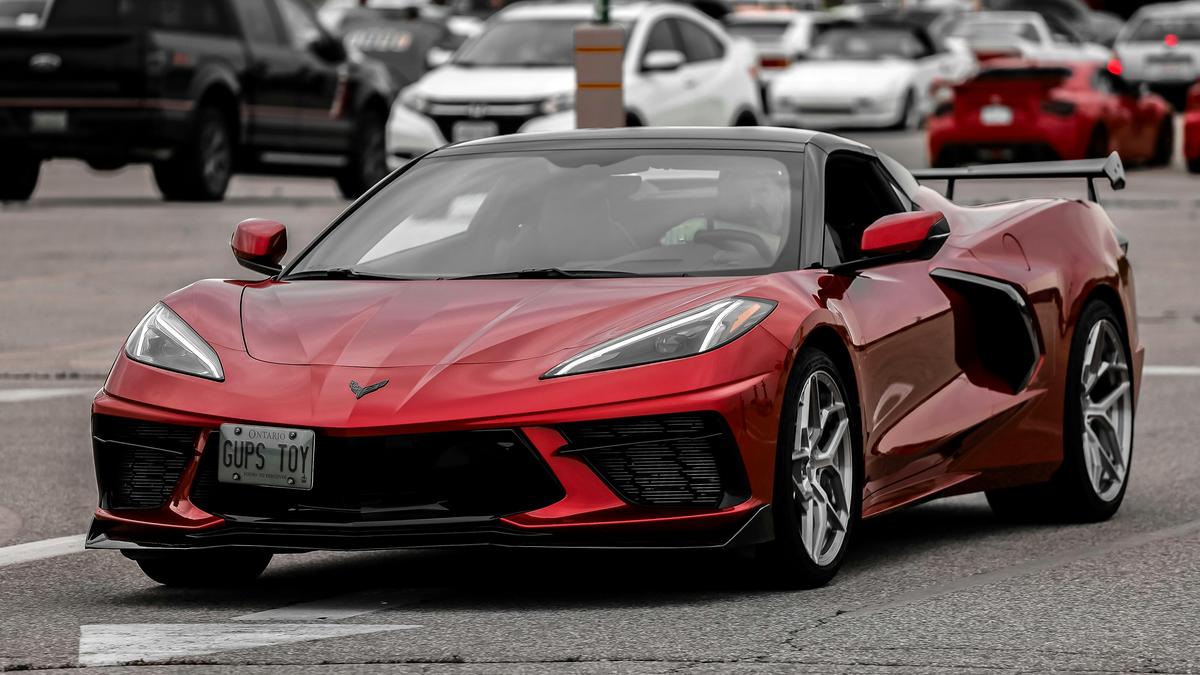A red Corvette C8 sports car with a personalized plate "GUPS TOY" is parked amidst other vehicles in a busy street.