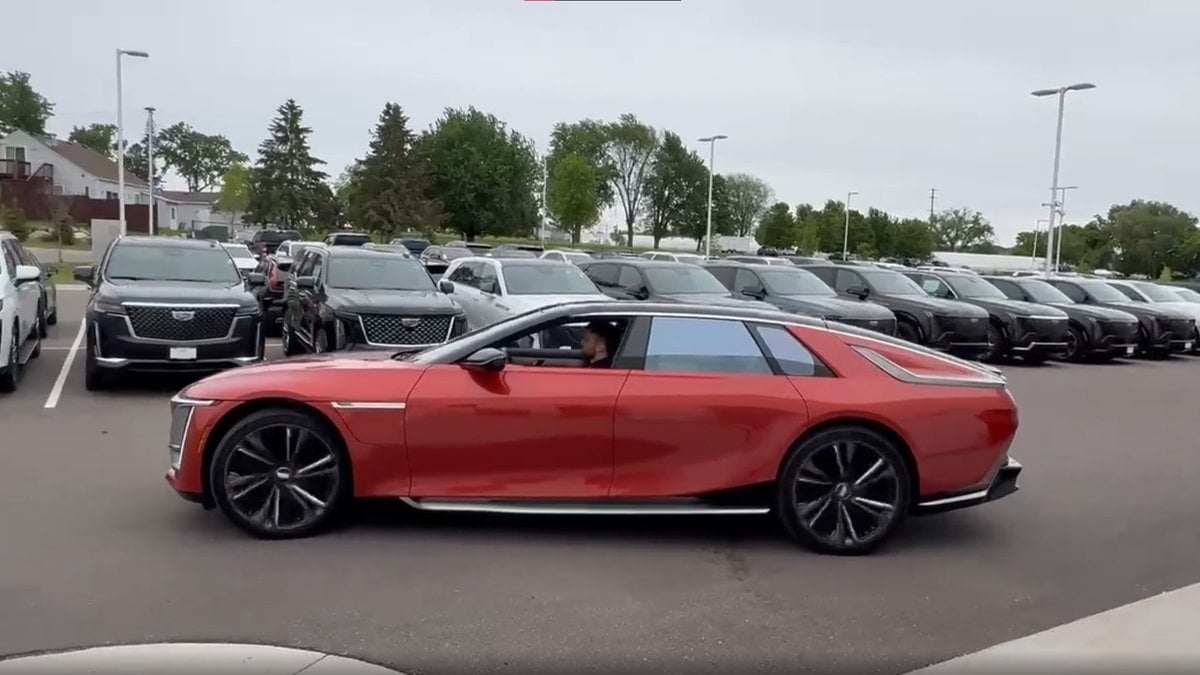 A 2026 Cadillac Celestiq, drives past rows of black SUVs in a parking lot, with trees and cloudy skies in the background.
