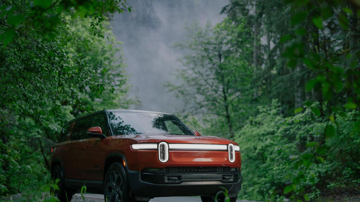 A Red 2025 Rivian R1S parked on a forested road, surrounded by lush green trees and misty mountains in the background.