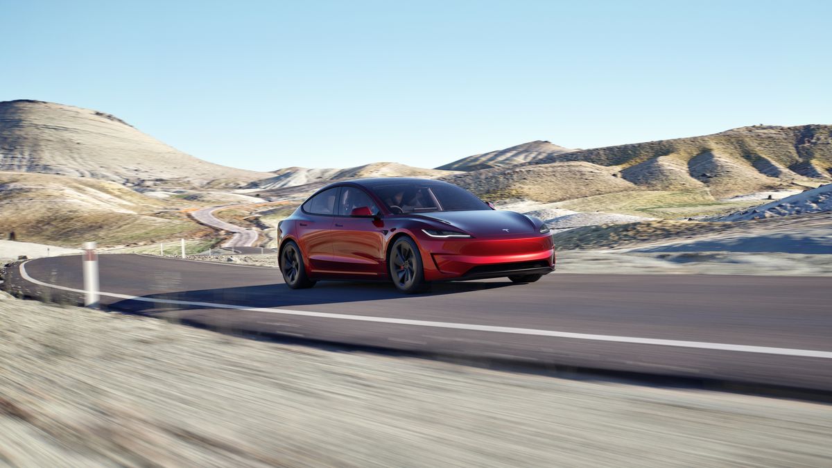 A red Tesla Model 3 speeds along a winding road, surrounded by rugged, hilly terrain under a clear blue sky.