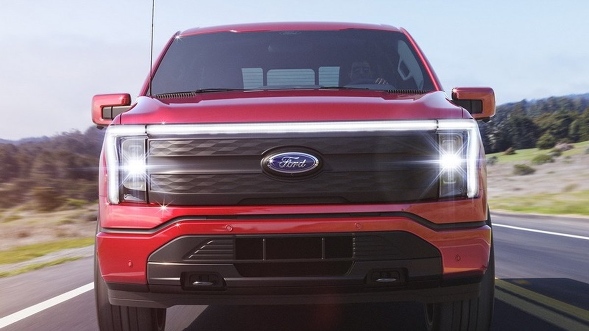 Front view of a red Ford electric truck on a scenic highway, showcasing its sleek design and bright headlights against a clear sky.