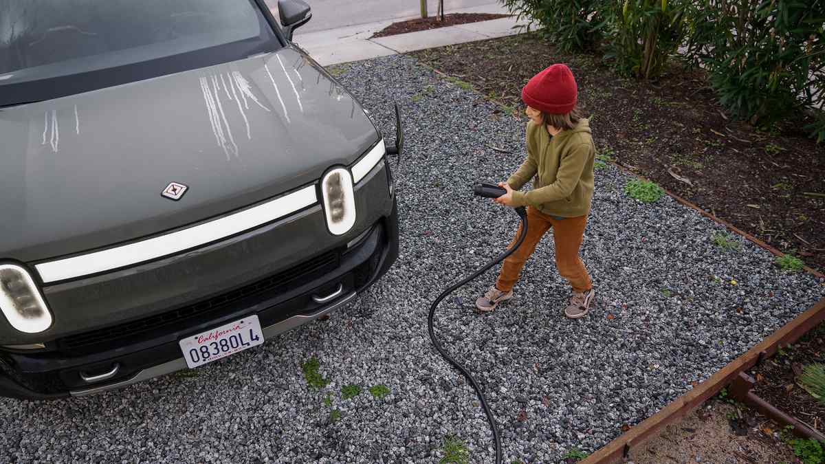 A child in a red hat plugs in a charging cable to a 2025 Rivian R1S parked on gravel, surrounded by greenery.