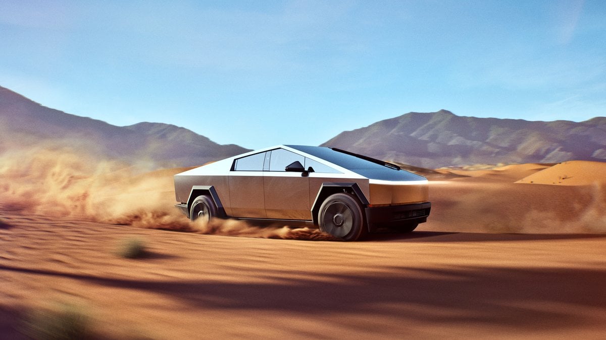 A futuristic silver Cybertruck speeds through a sandy desert landscape, kicking up dust against a backdrop of mountains and clear blue skies.