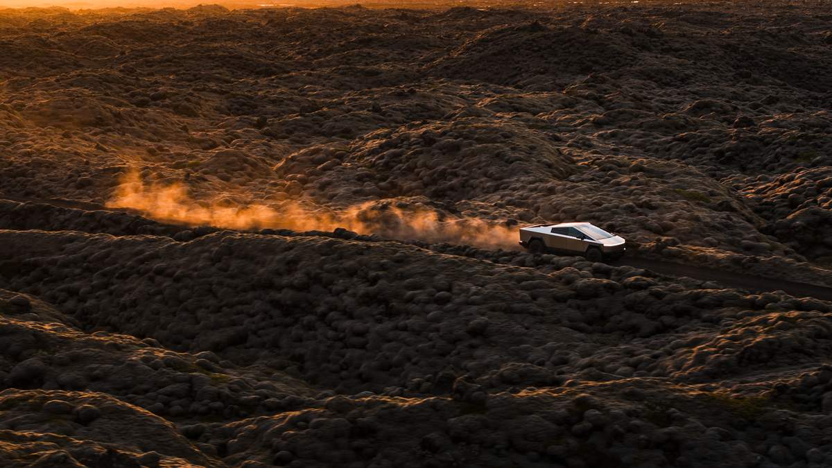 A Tesla Cybertruck drives over rugged, volcanic terrain at sunset, leaving a trail of dust in its wake against a dramatic landscape.