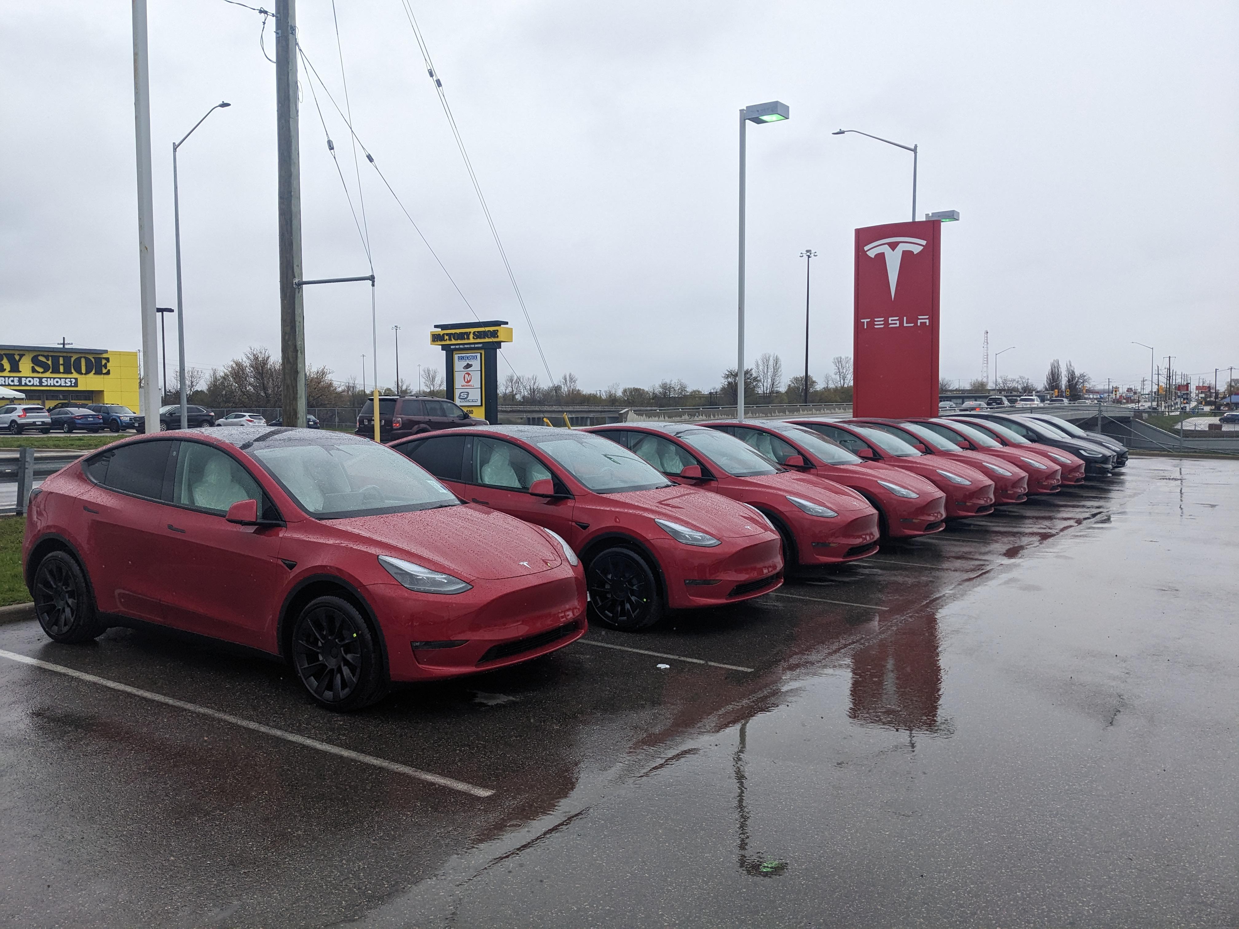 A row of red Tesla Model Y vehicles parked under gray skies, with a prominent Tesla sign in the background.