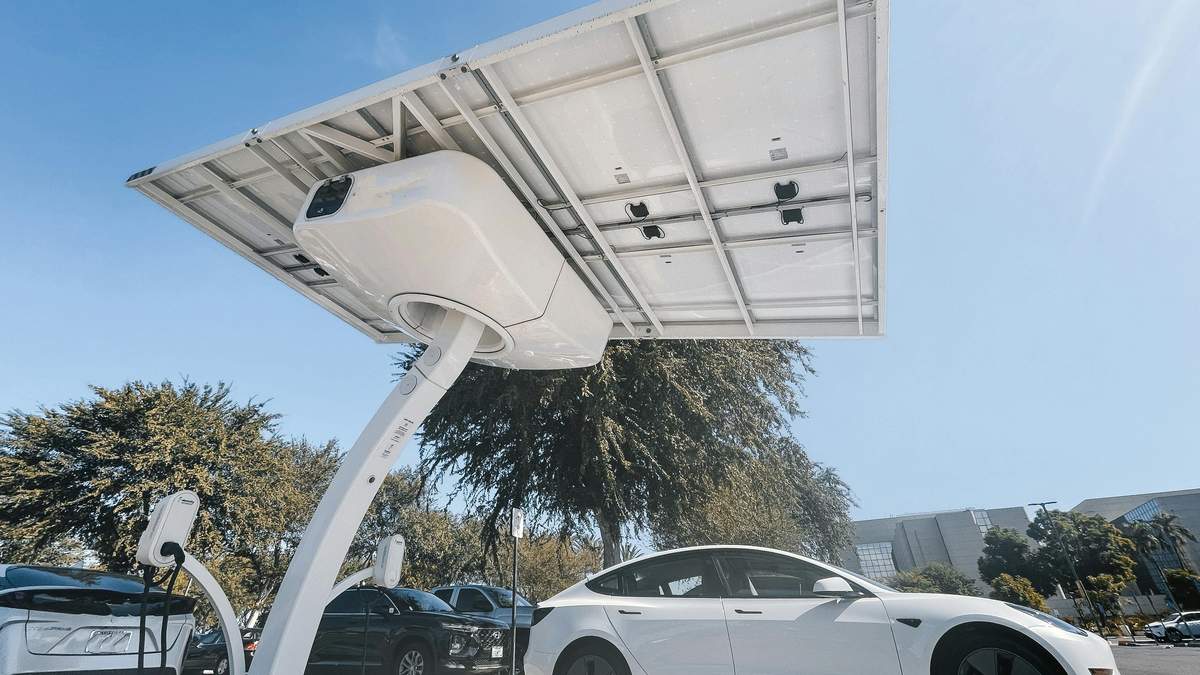 A white Tesla Model 3 parked beneath a charging station, surrounded by trees and clear blue sky.