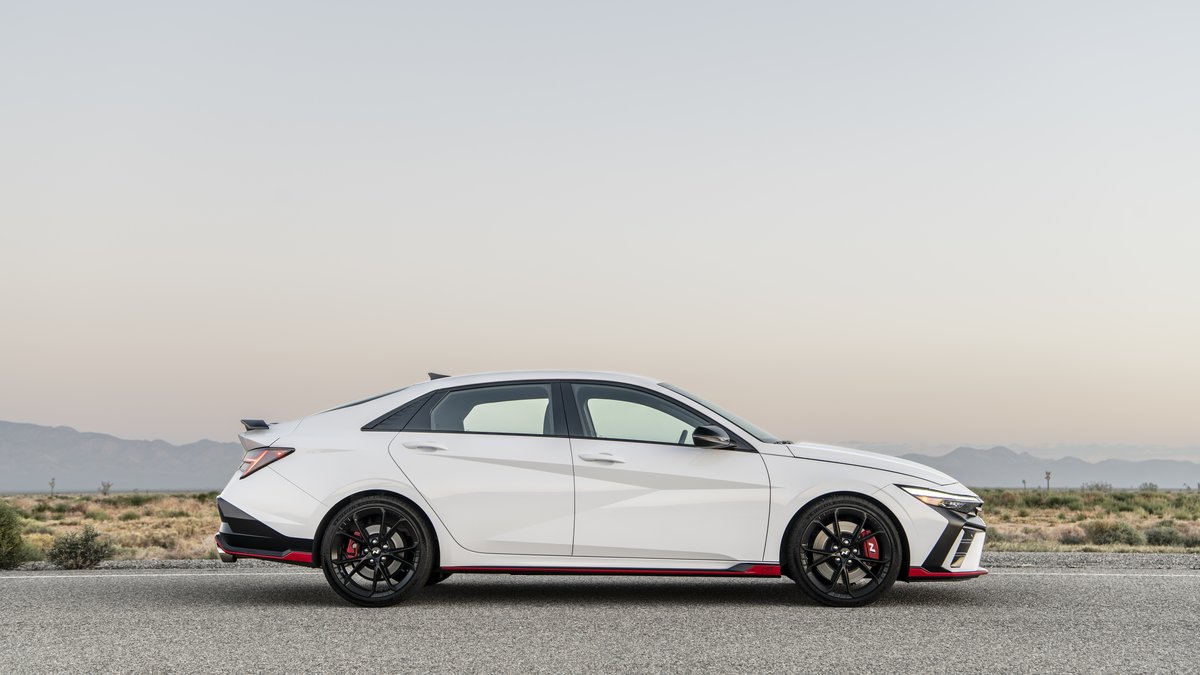 A white, 2025 Hyundai Elantra N with black wheels and red accents is parked on a road, with a desert landscape and mountains in the background.