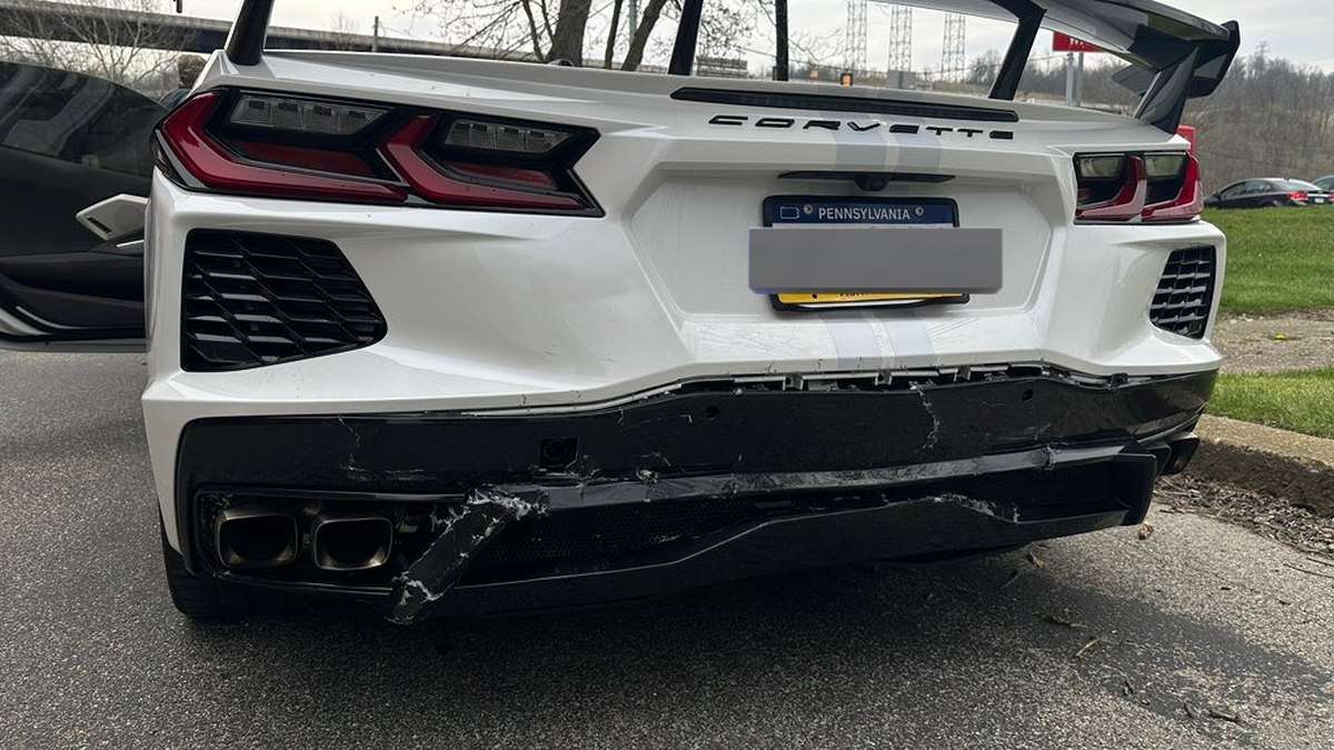 A white Corvette with a damaged rear bumper and exhaust, parked in a lot, showcasing a prominent wing and broken bodywork.