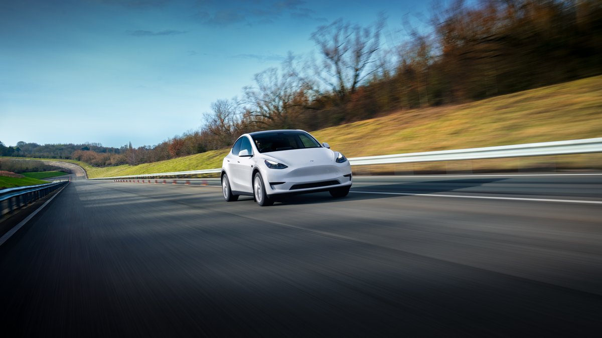 A white electric car speeds along a curved highway, surrounded by green grass and trees under a clear blue sky.