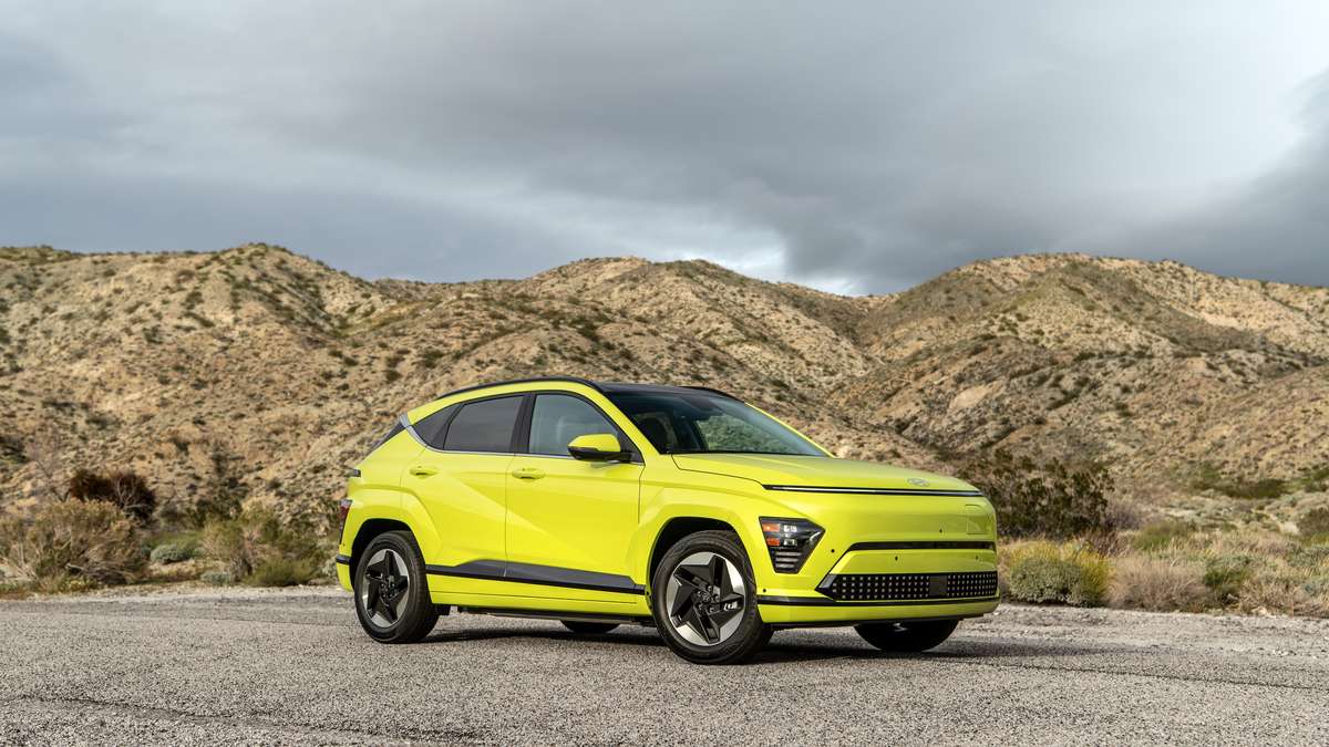A bright green 2025 Hyundai Kona EV parked on a gravel road with rugged mountains in the background, under a partly cloudy sky.