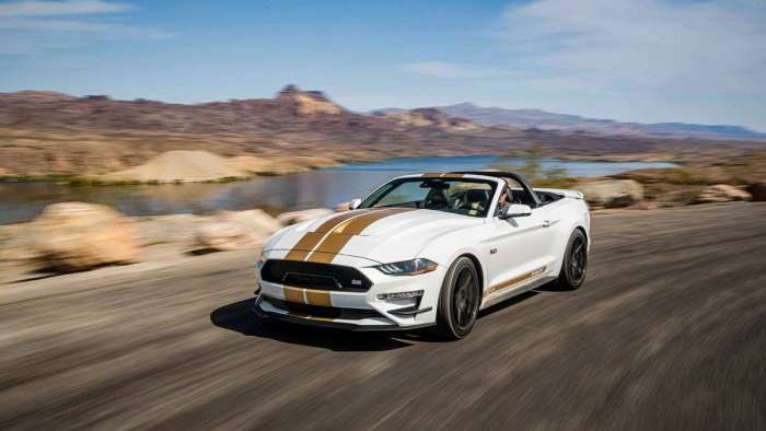 Image of a white convertible Shelby GT-H with gold racing stripes driving on a canyon road.